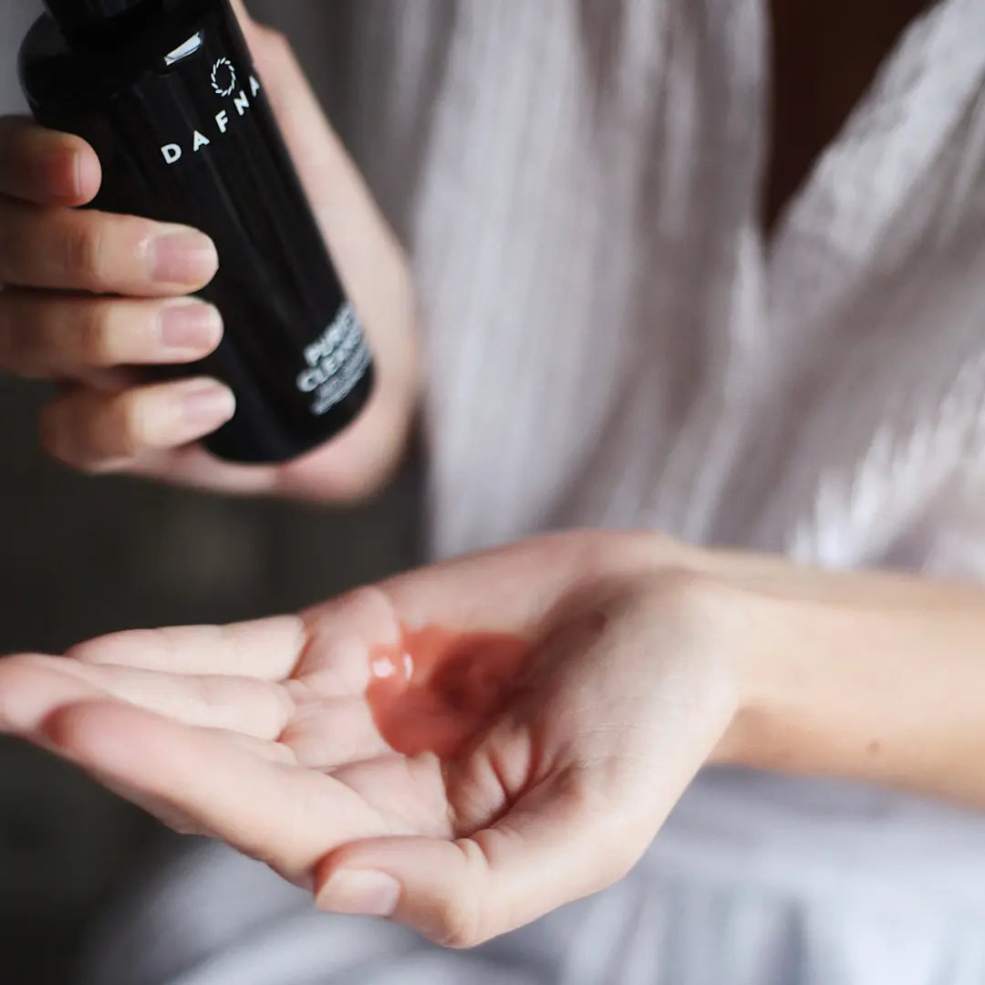 Rich dark red color of Purify cleanser, on a hand. The other hand is holding the bottle.